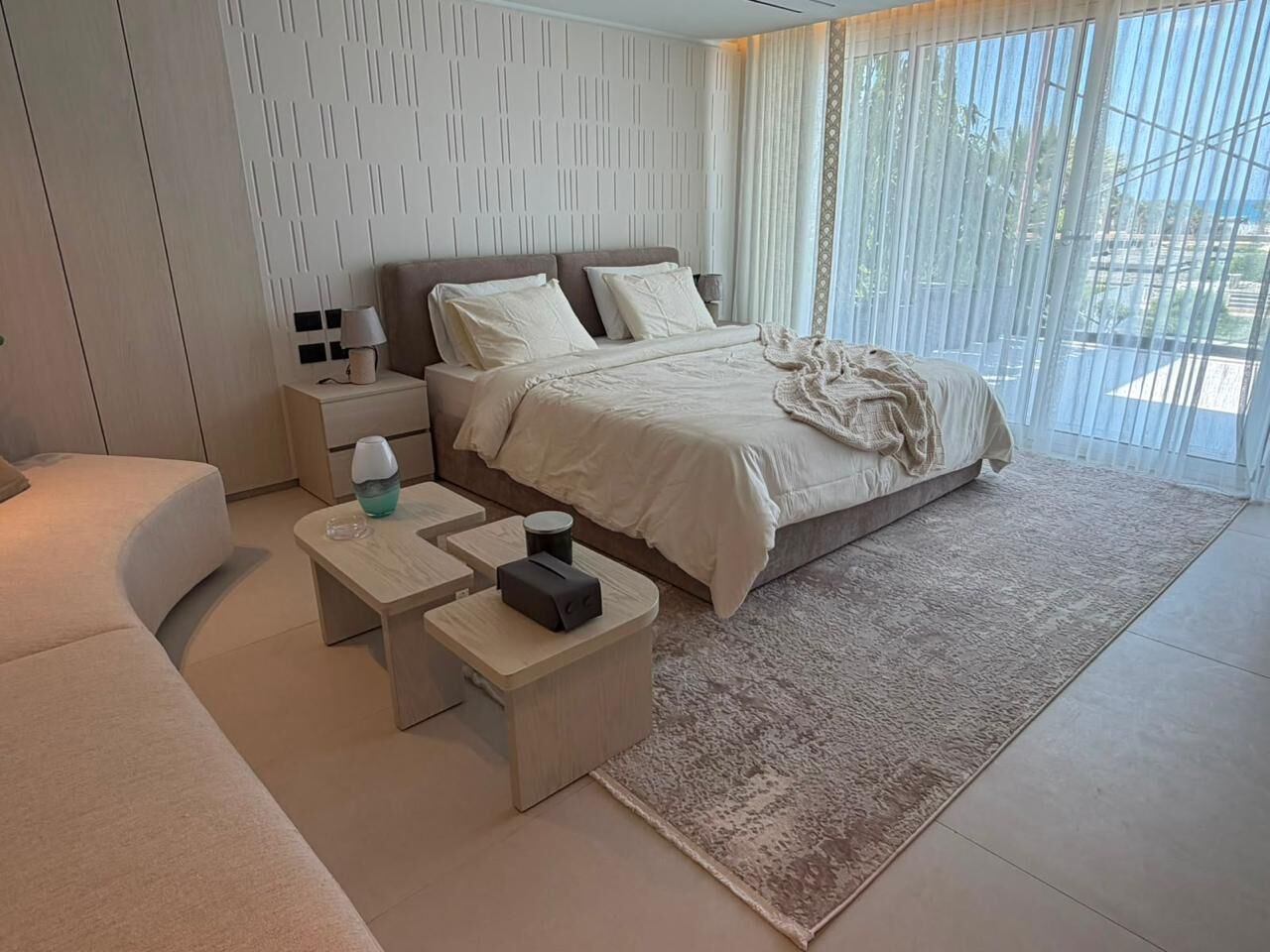 Bed with soft bedding on a grey and cream rug, with a nightstand and curtains