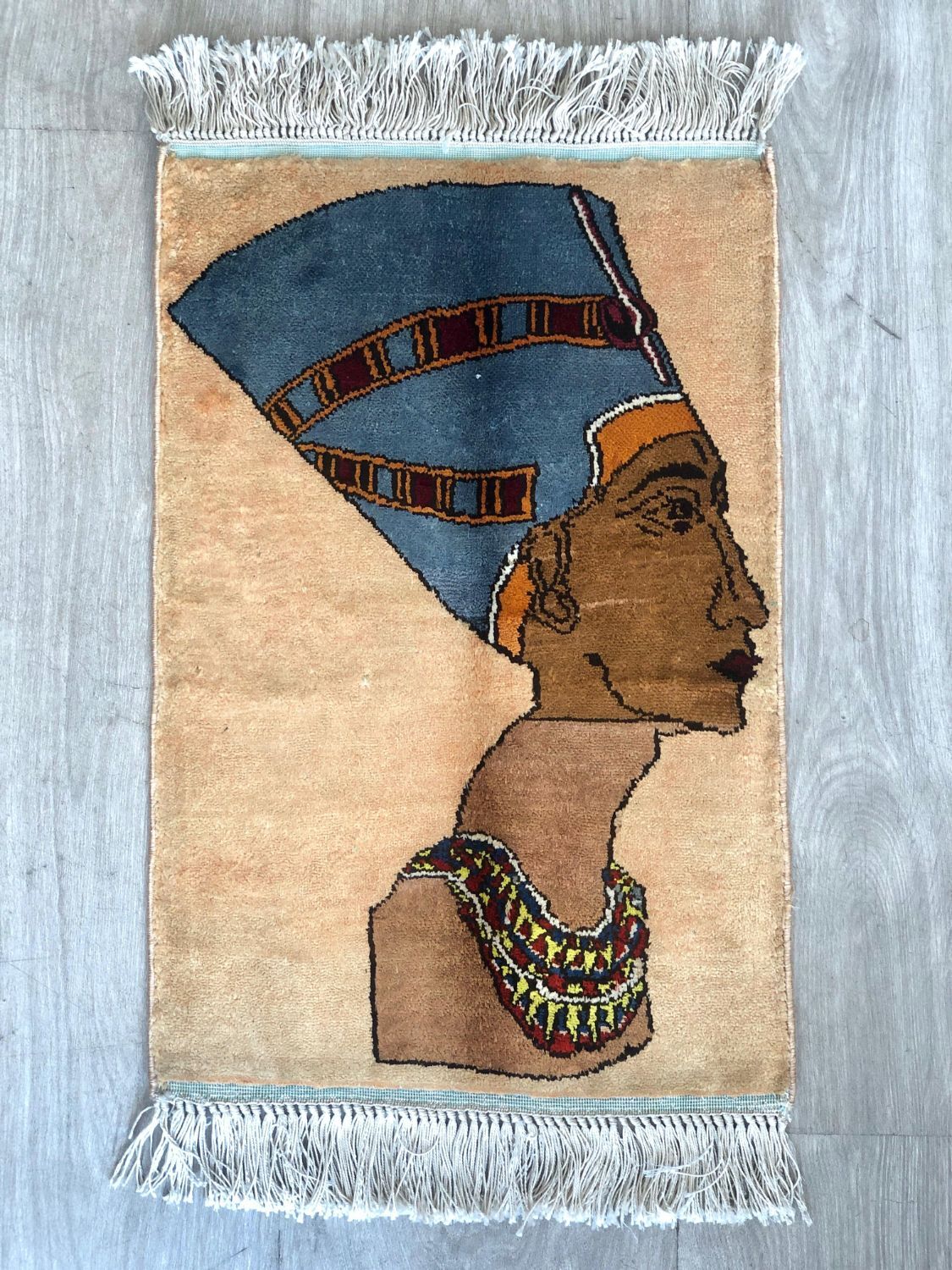 Small pictorial rug showing a profile bust of a queen in a tall blue crown with a broad jewelled collar, on a peach field