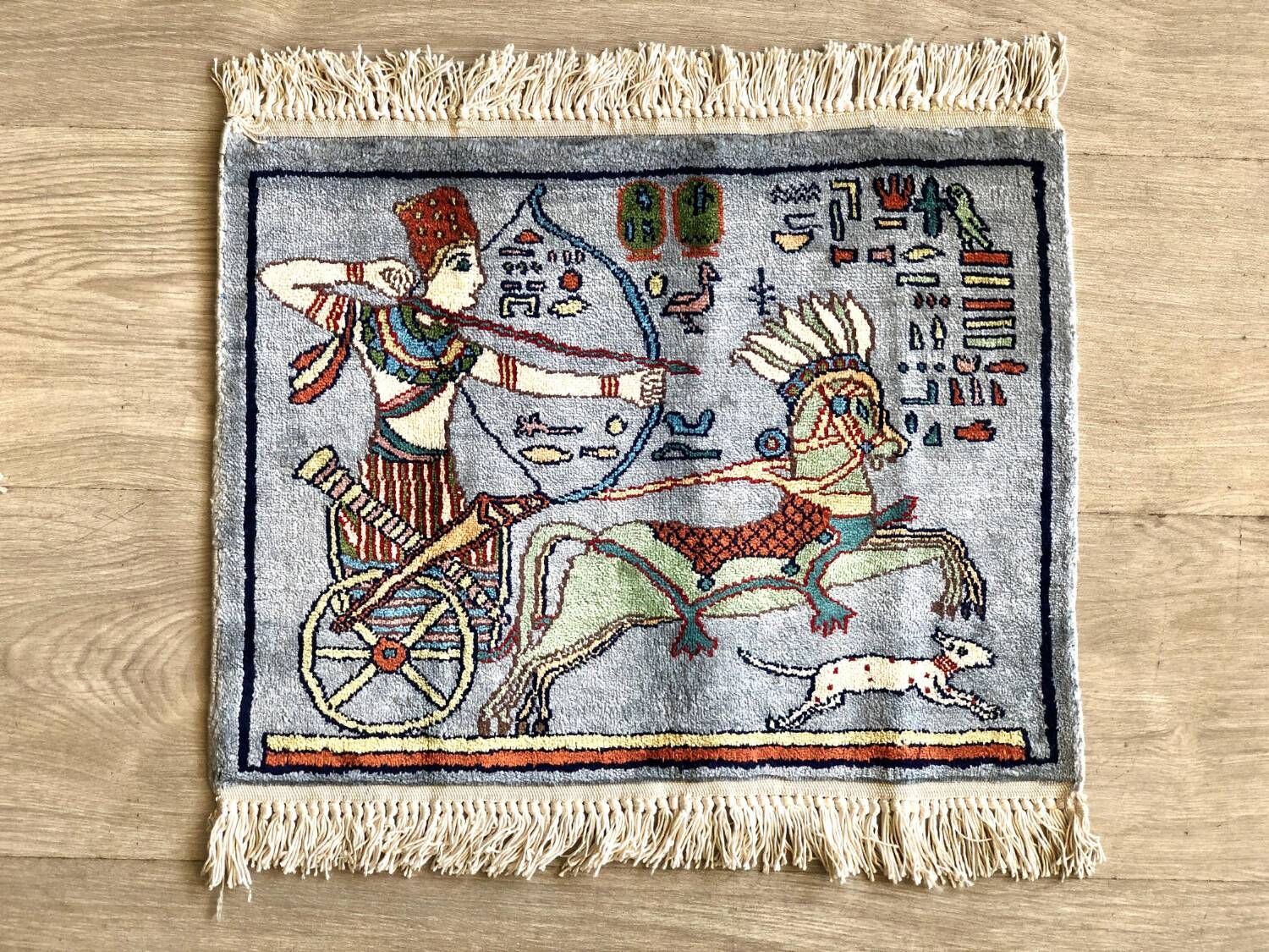 Small pictorial rug showing an archer in a horse-drawn chariot with a running dog and hieroglyph-style symbols, on a blue-grey field