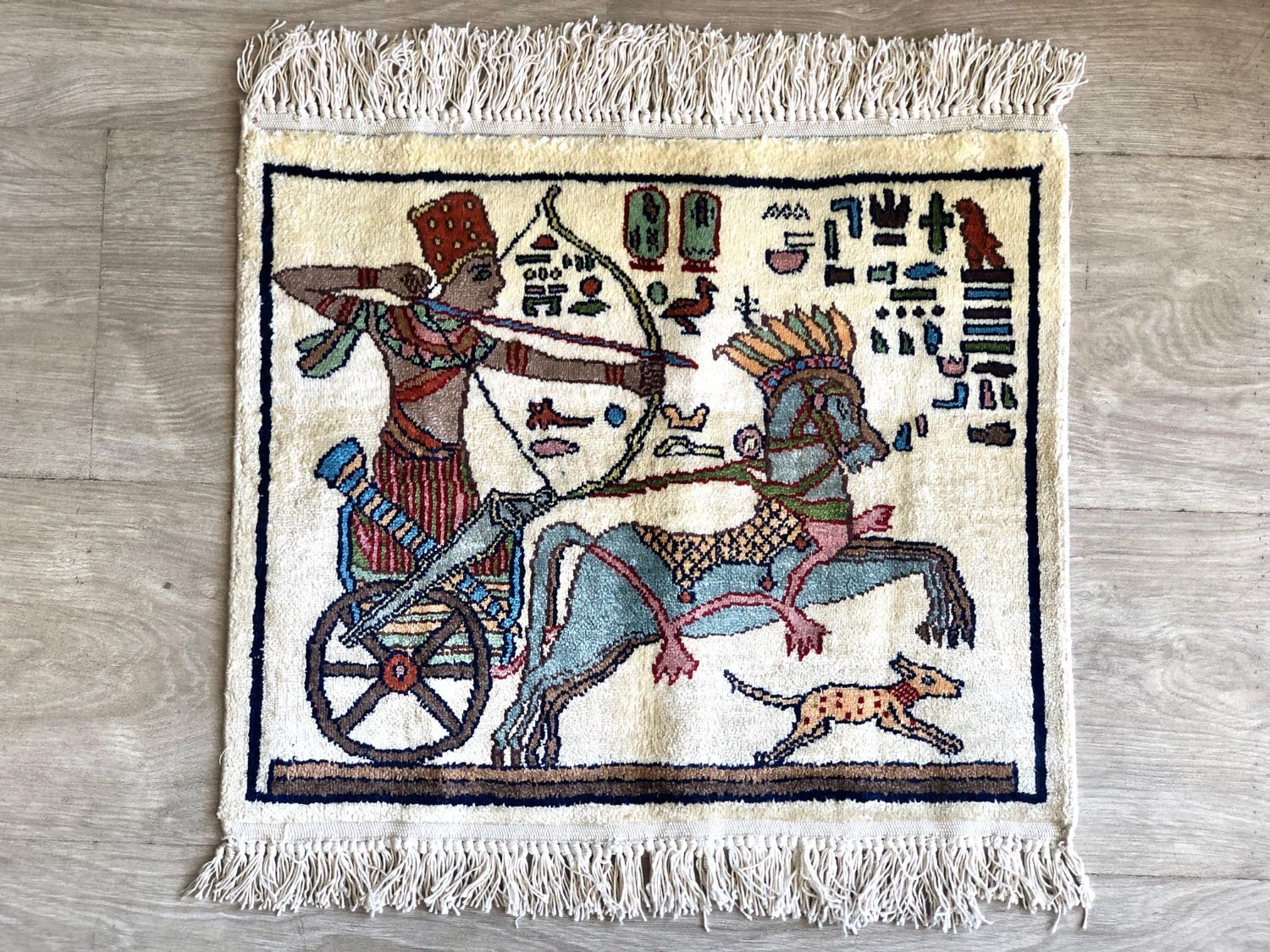 Small pictorial rug showing an archer in a horse-drawn chariot with a running dog and hieroglyph-style symbols, on a cream field
